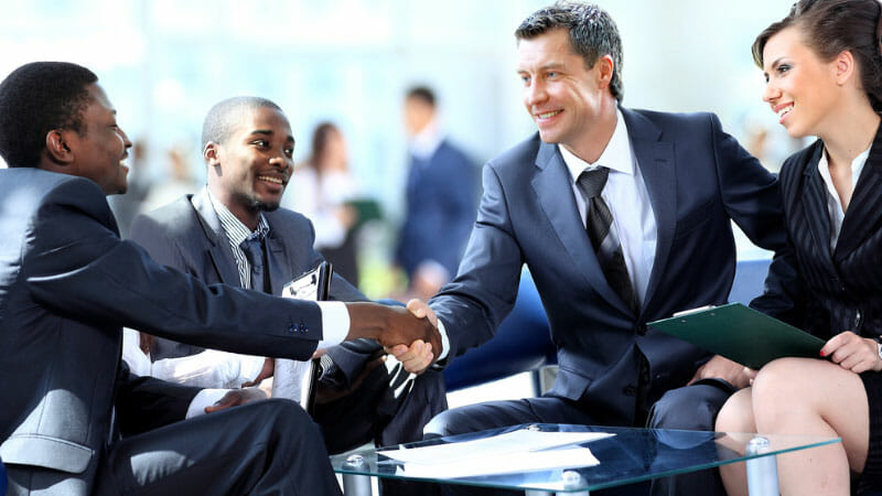 Four business professionals, three men and one woman, sit together in an office, with two men in the group shaking hands and others smiling and holding documents.