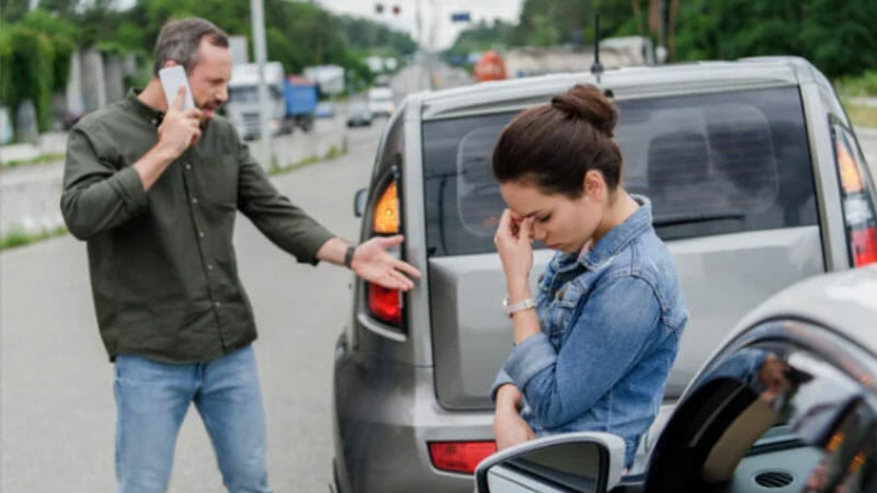 A man talks on the phone and gestures towards a woman who looks upset, standing near two cars that appear to have been in a minor accident on a road.