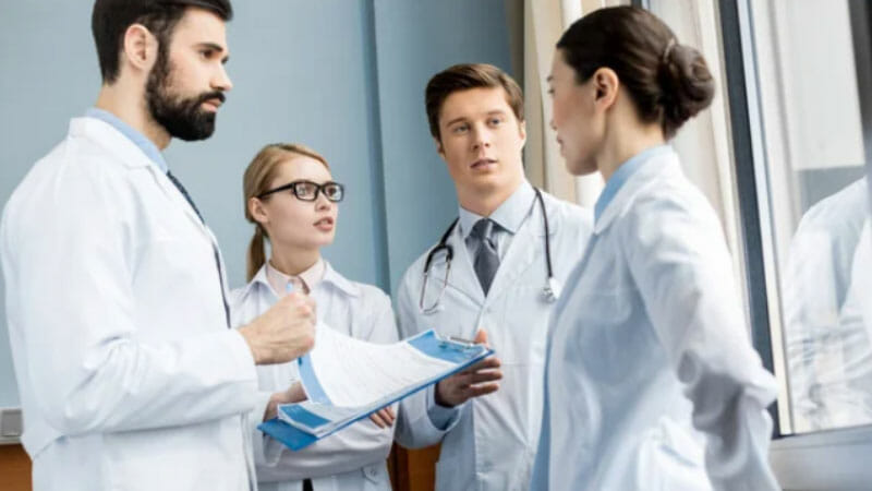 Four doctors in white coats stand together indoors, discussing documents and looking attentive.