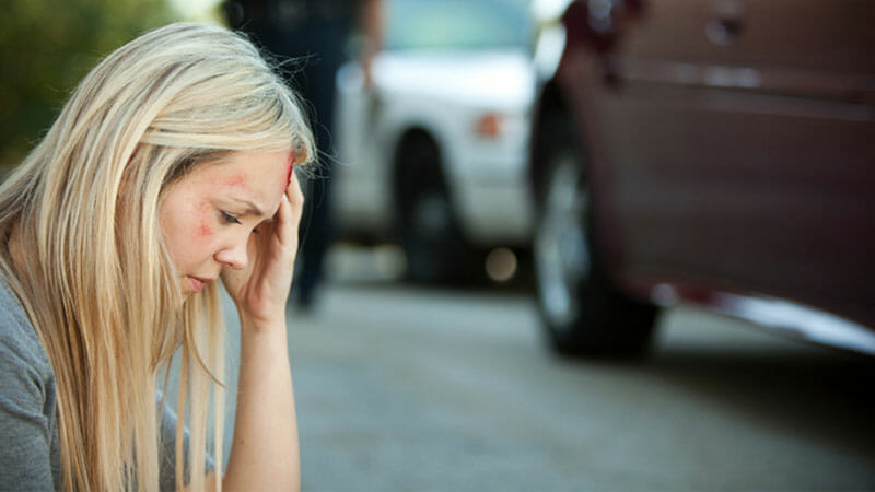 A woman with facial injuries sits on the ground beside a car, holding her head, with emergency responders and vehicles in the background.