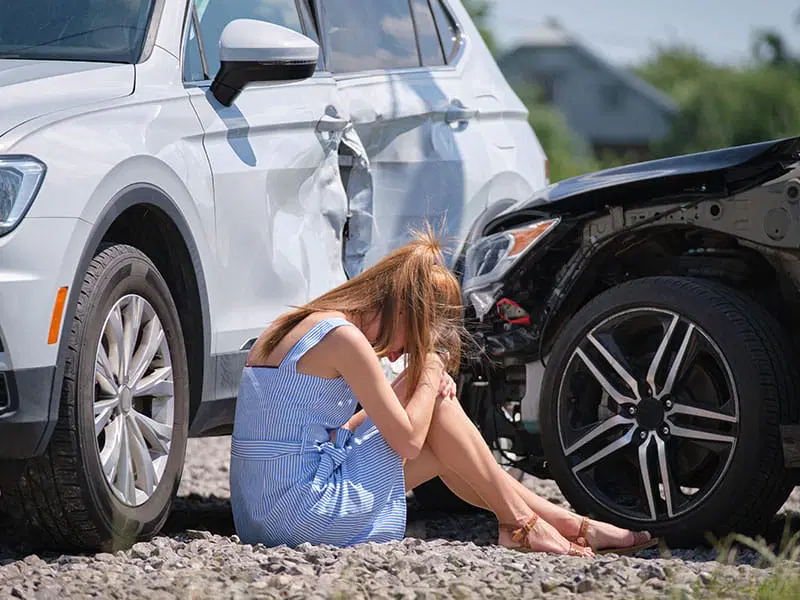 A woman in a blue dress sits on the ground, holding her head, next to two damaged cars involved in a collision.