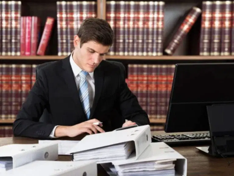 A man in a suit sits at a desk reviewing documents, with stacks of binders, a computer, and bookshelves in the background.