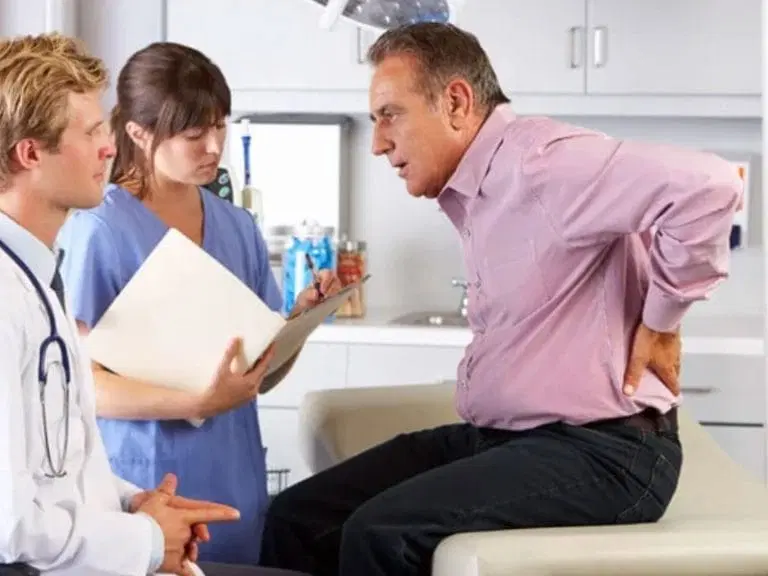 An older man sitting on an exam table holds his lower back while talking to a doctor and nurse in a medical office.