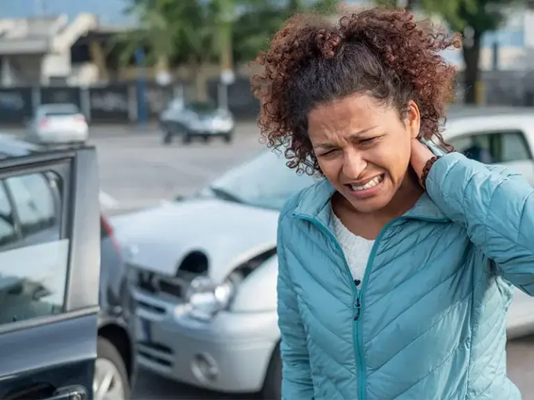 A woman in a blue jacket holds her neck in pain near damaged cars after a traffic accident in a parking lot.
