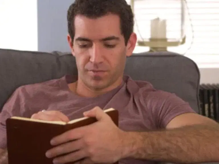 A man sitting on a couch, wearing a maroon t-shirt, reading a brown notebook.