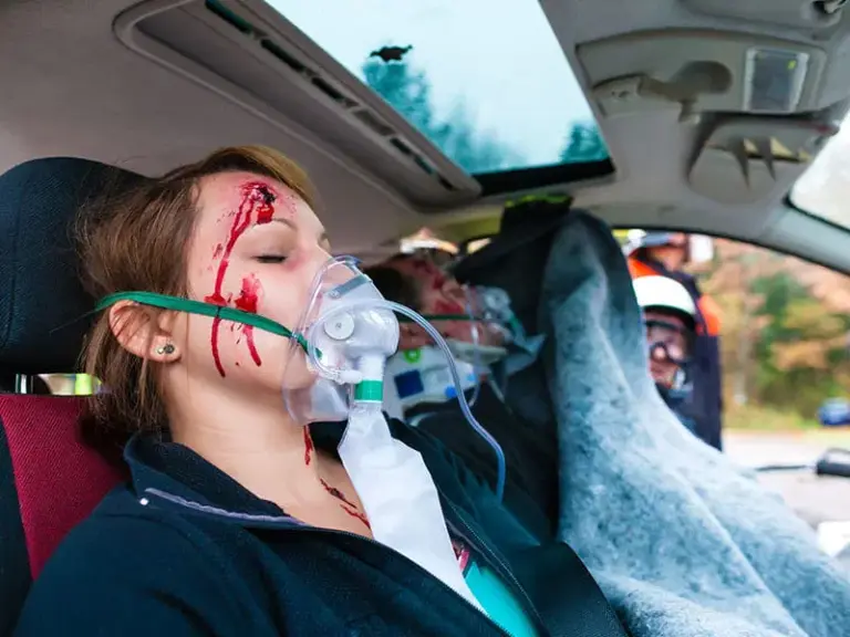 A woman with facial injuries and an oxygen mask sits in a car, with emergency responders attending to her and another injured passenger in the background.