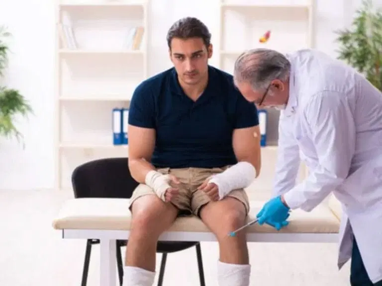 A doctor in a white coat administers an injection to a seated man with bandaged arms and legs in a medical office.
