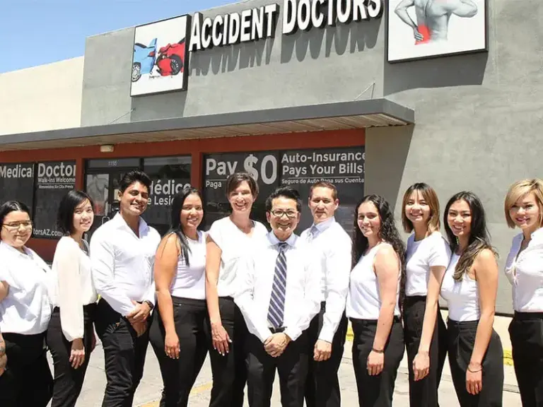 A group of ten people stand in front of a building with a sign that reads "Accident Doctors." They are dressed in coordinated white tops and dark bottoms, smiling at the camera.