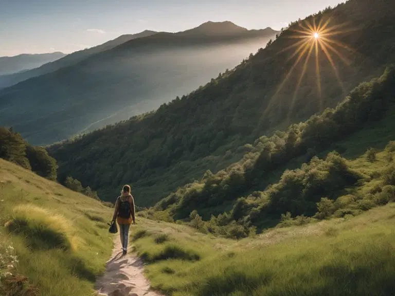 A person with a backpack walks along a grassy mountain trail at sunrise, with sunlight streaming over a forested ridge in the background.