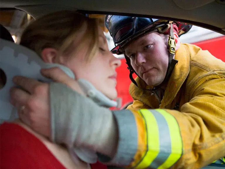 A firefighter in uniform fits a neck brace on a woman sitting in a car, assisting her after an apparent accident.
