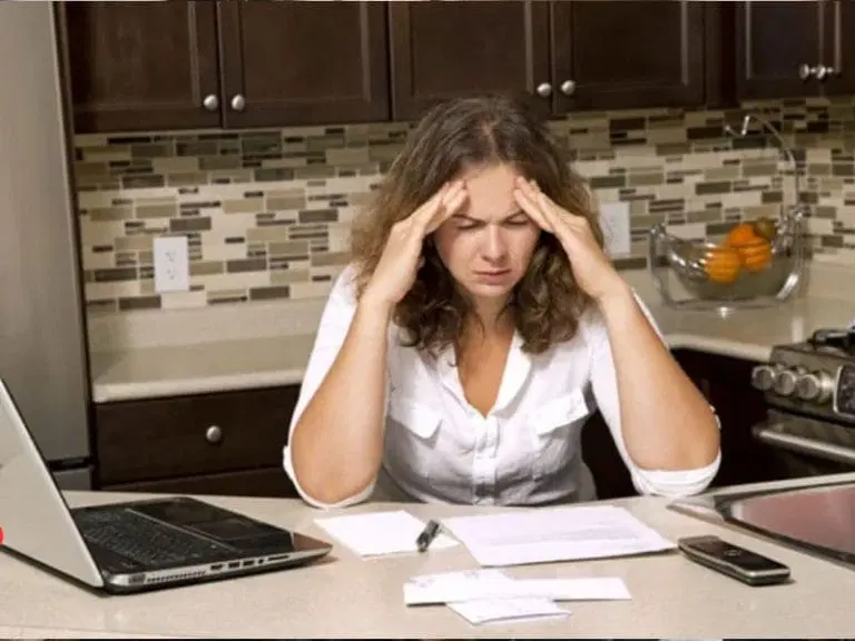 A woman sits at a kitchen counter with her head in her hands, looking stressed while reviewing papers next to a laptop and phone.