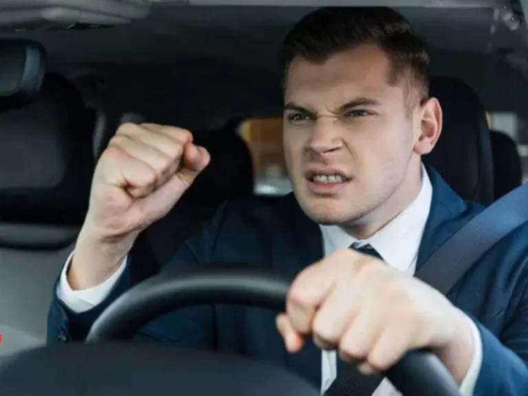 A man in a suit sits in the driver’s seat of a car, gripping the steering wheel with one hand and raising his clenched fist in frustration—highlighting the importance of Safe Driving Tips to manage road rage.