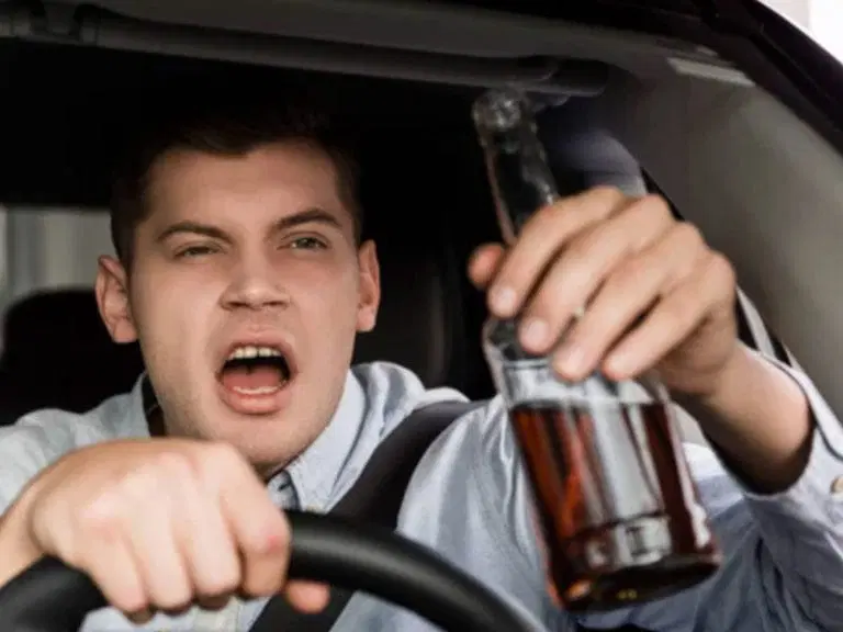 A man is sitting in the driver’s seat of a car, gripping the steering wheel with one hand and holding a bottle of alcohol with the other—an example that highlights the importance of following safe driving tips. His mouth is open.