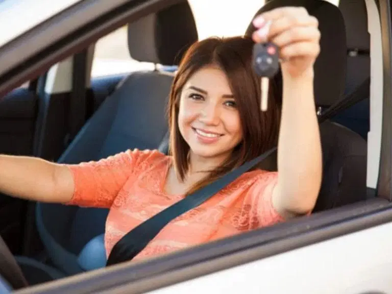 Woman sitting in the driver's seat of a car, wearing a seatbelt, smiling, and holding up a car key—ready to hit the road with Safe Driving Tips in mind.