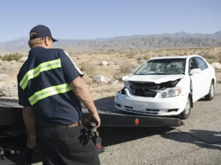 A tow truck operator prepares to load a white car with front-end damage onto a flatbed in a desert area, reminding us of the importance of following Safe Driving Tips.