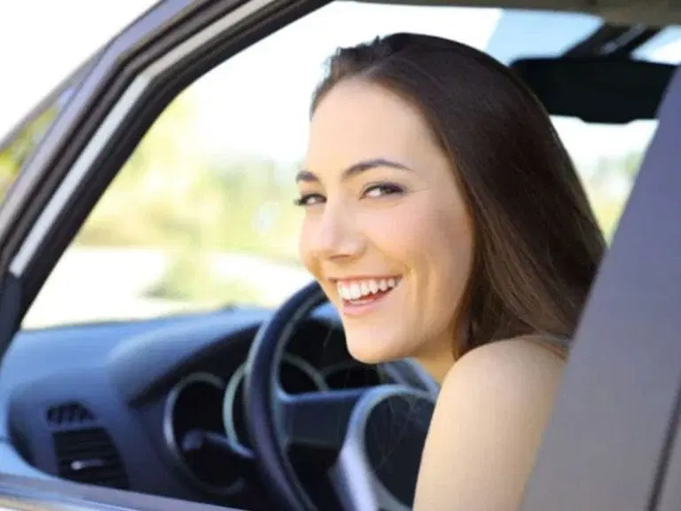 A woman with long brown hair smiles while sitting in the driver's seat of a car with the window open, ready to hit the road and follow safe driving tips.