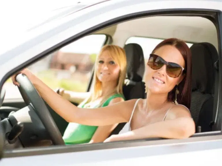 Two women sitting in the front seats of a car, one driving and both smiling at the camera, perfectly embodying Safe Driving Tips by wearing seatbelts and staying attentive on the road.