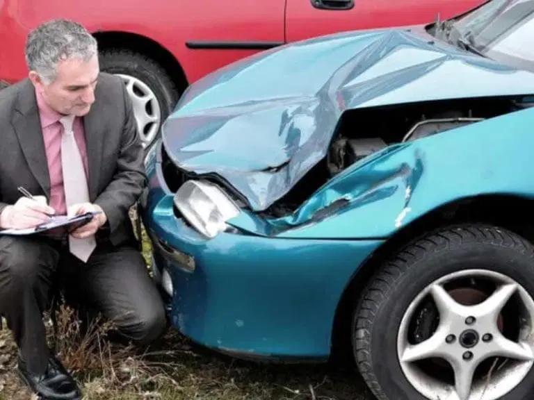 A man in a suit examines and takes notes on a blue car with significant front-end damage, parked next to a red car.