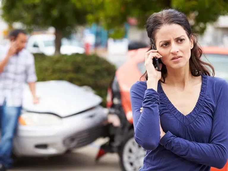 A woman stands in front of a car accident, talking on her phone, while a man in the background also makes a phone call next to damaged vehicles.