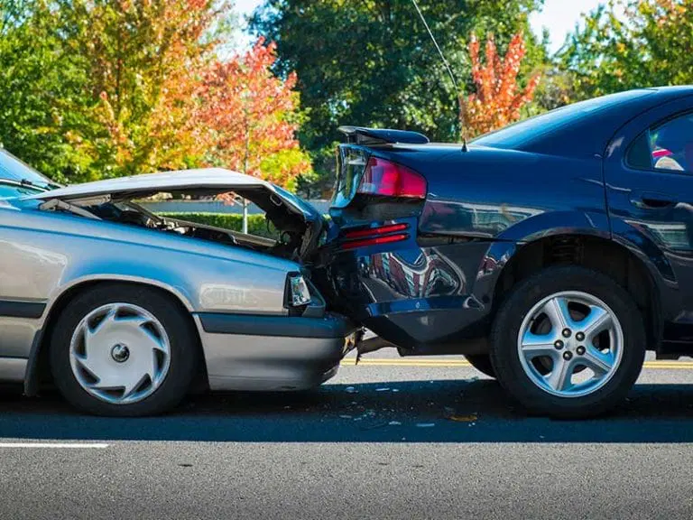 A silver car has rear-ended a black car on a road, causing visible damage to both vehicles. Trees with fall foliage are in the background.