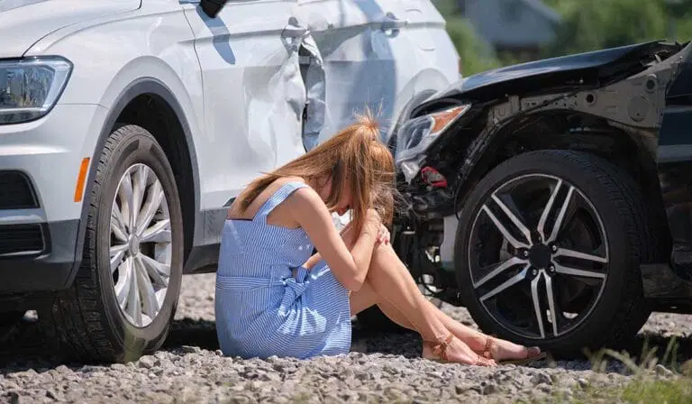 A woman in a blue dress sits on gravel with her head in her hands near two cars that appear to have collided, showing visible damage.