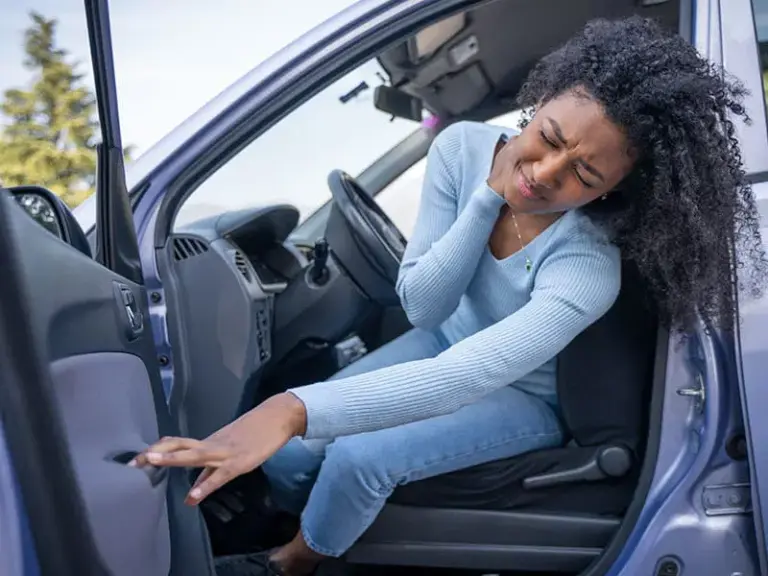 A woman sitting in a car holds her neck in pain while reaching for the door handle to get out.