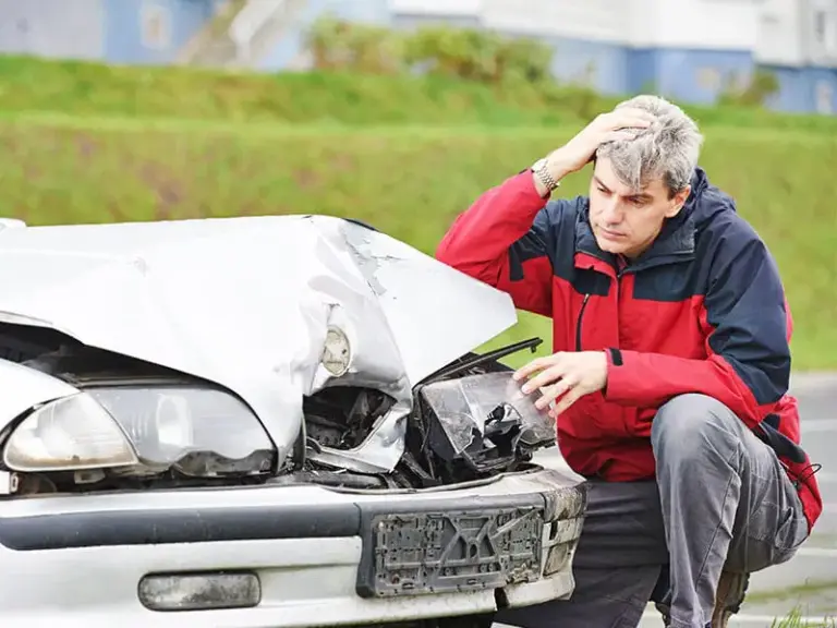 A man in a red jacket examines the damaged front end of a white car after an apparent crash, looking concerned and holding his head.