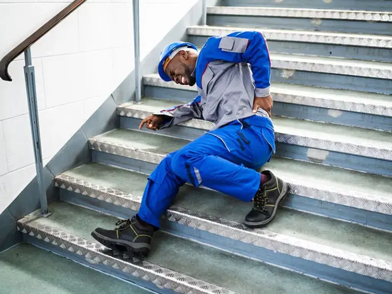 A worker in blue coveralls and a hard hat is lying on a staircase, holding his lower back and appearing to be in pain after a fall.