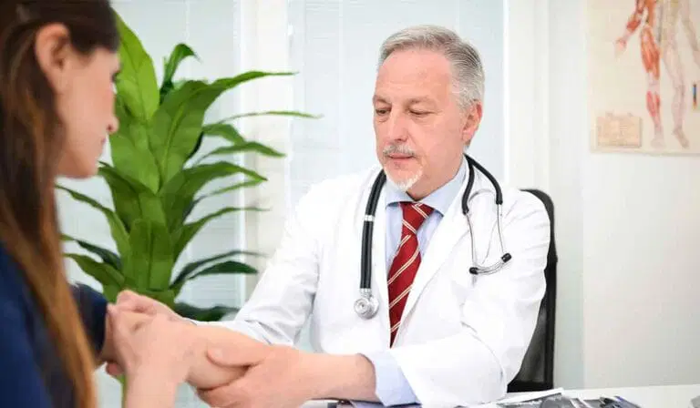 A doctor in a white coat examines a patient's arm in a medical office with a plant and anatomical chart in the background.