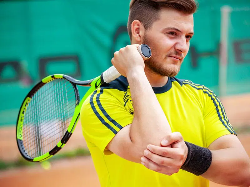 A man in a yellow shirt holds his tennis racket over his shoulder and touches his elbow, appearing to feel discomfort on a tennis court.