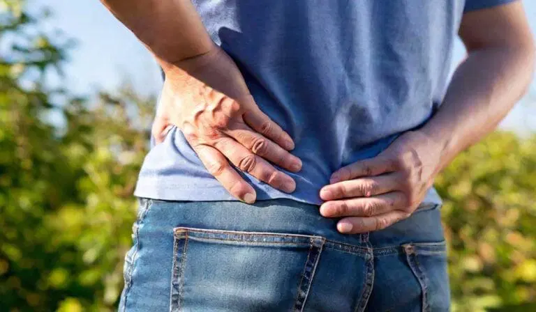 A person wearing jeans and a blue shirt places their hands on their lower back and hip, appearing to experience discomfort or pain.