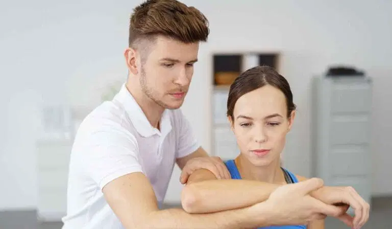 A physical therapist assists a woman by gently stretching her arm in a clinical setting.