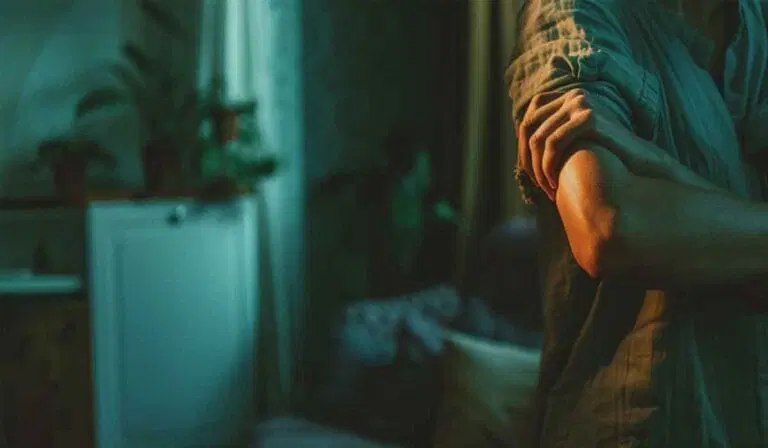 A person stands indoors with arms crossed, partially visible, in a dimly lit room with plants and a closed door in the background.