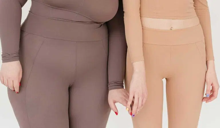 Two people wearing fitted, long-sleeve tops and high-waisted leggings in brown and beige tones, standing side by side against a neutral background.