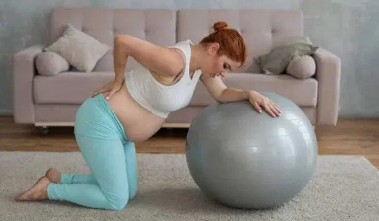 Pregnant woman kneeling on a rug, holding her lower back with one hand and leaning on an exercise ball with the other, in a living room setting.