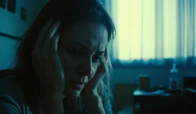 A woman sits indoors with her hands on her head, looking distressed, as light filters through a window in the background.