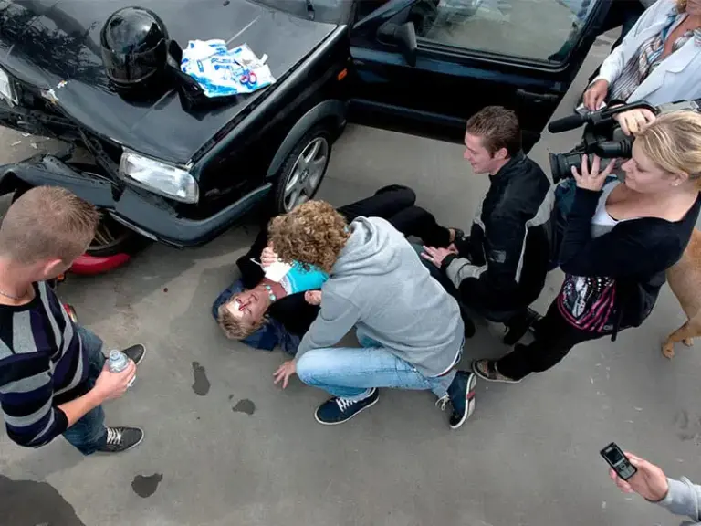 A group of people assist an injured person lying on the ground near a damaged car, while others observe and take photos.