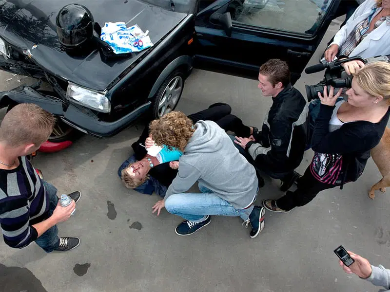 A group of people assist an injured person lying on the ground near a damaged car, while others observe and take photos.
