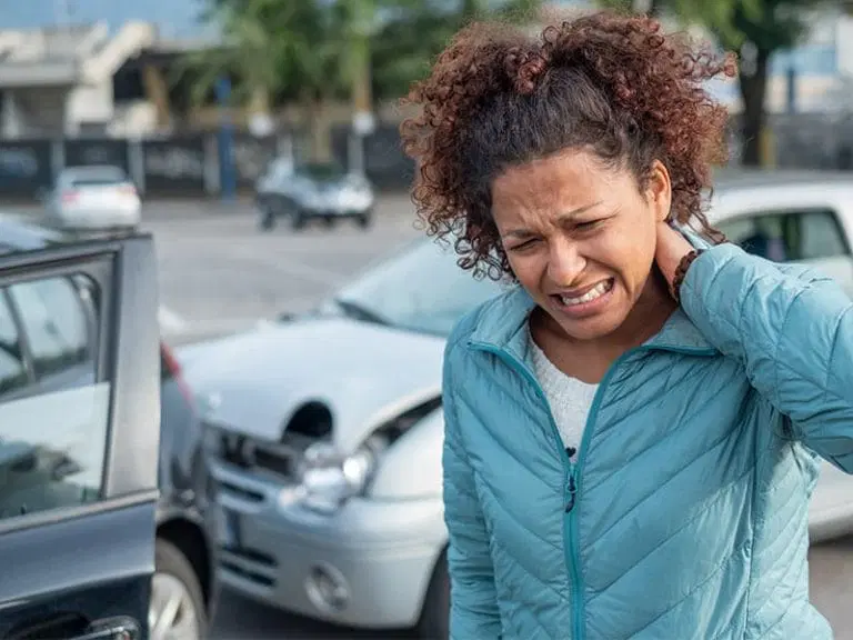 A woman in a blue jacket holds her neck in pain next to damaged cars after a traffic accident in a parking lot.