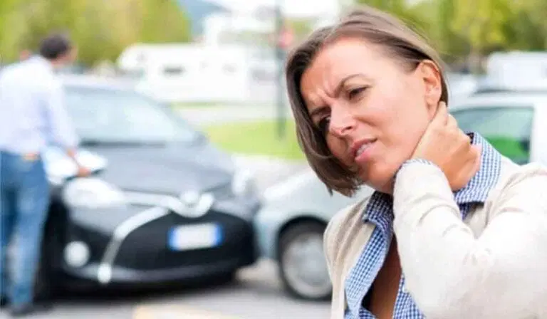 A woman holds her neck in apparent pain, standing in front of two cars that appear to have been in a minor accident. Another person is near the vehicles.