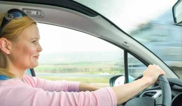 A woman with blonde hair drives a car during the day, holding the steering wheel with both hands and looking straight ahead. The background shows a blurred landscape and a passing vehicle.