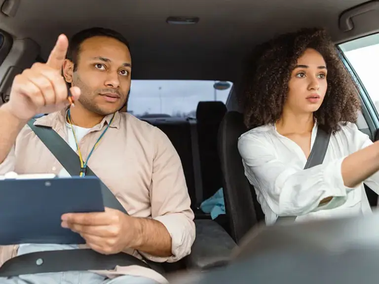 A man holding a clipboard sits in the passenger seat, pointing, while a woman drives a car, both wearing seatbelts.