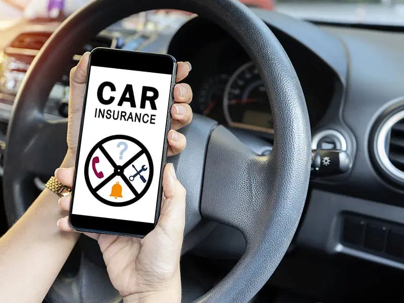 A person holds a smartphone displaying a car insurance app while sitting in the driver’s seat of a car.