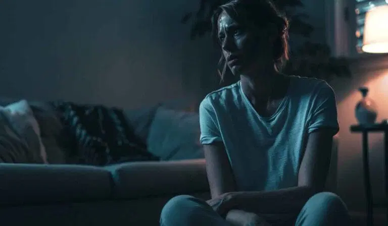 A woman sits on the floor in a dimly lit room, appearing thoughtful or worried. A couch and a lamp are visible in the background.