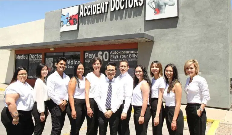 A group of eleven people in business attire pose in front of an "Accident Doctors" clinic building on a sunny day.