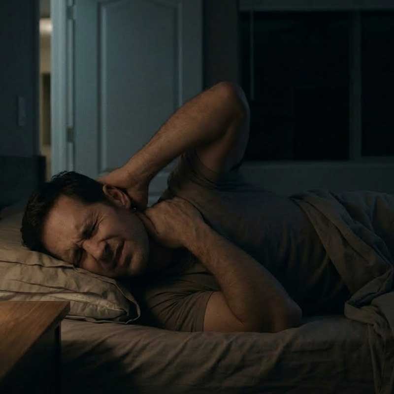A man lies in bed at night, grimacing in pain as he holds his neck and upper back with both hands.