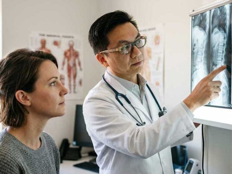A doctor explains X-ray results to a patient in a medical office, pointing at images displayed on a lightbox.