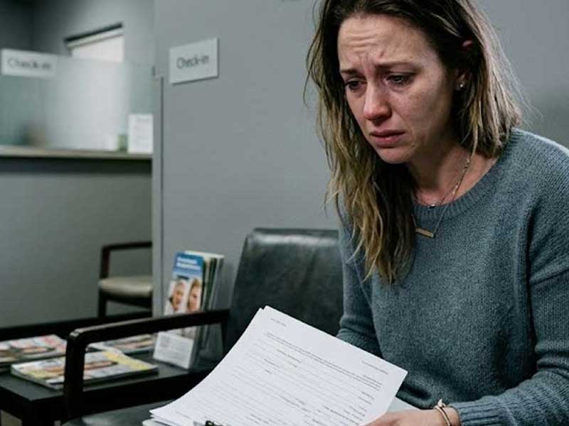 A woman sits in a waiting room, visibly upset, holding paperwork. Signs reading "Check-In" are visible in the background.