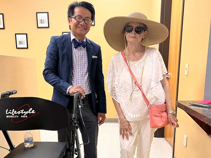 A man in a suit stands next to an older woman wearing a large sun hat and sunglasses. A mobility aid labeled "lifestyle" is nearby. They are indoors in a room with framed art on the wall.