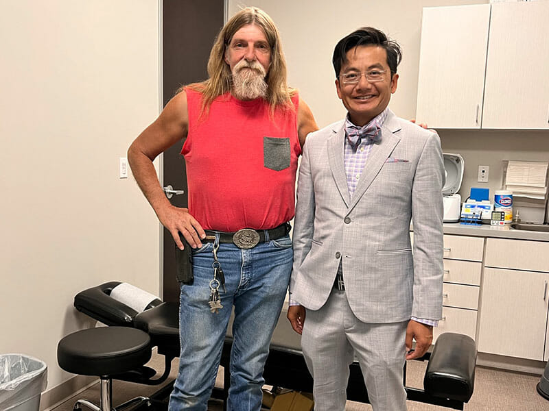 Two men stand in a medical office in Mesa; one wears a red sleeveless shirt and jeans, the other sports a light gray suit and bow tie. Medical equipment and supplies are visible, suggesting an experienced Accident Doctor is present.